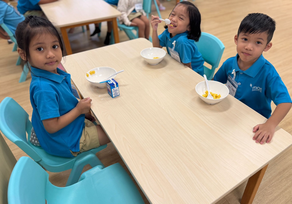 Three children in blue shirts sit at a table with bowls and spoons, eating cereal with milk.